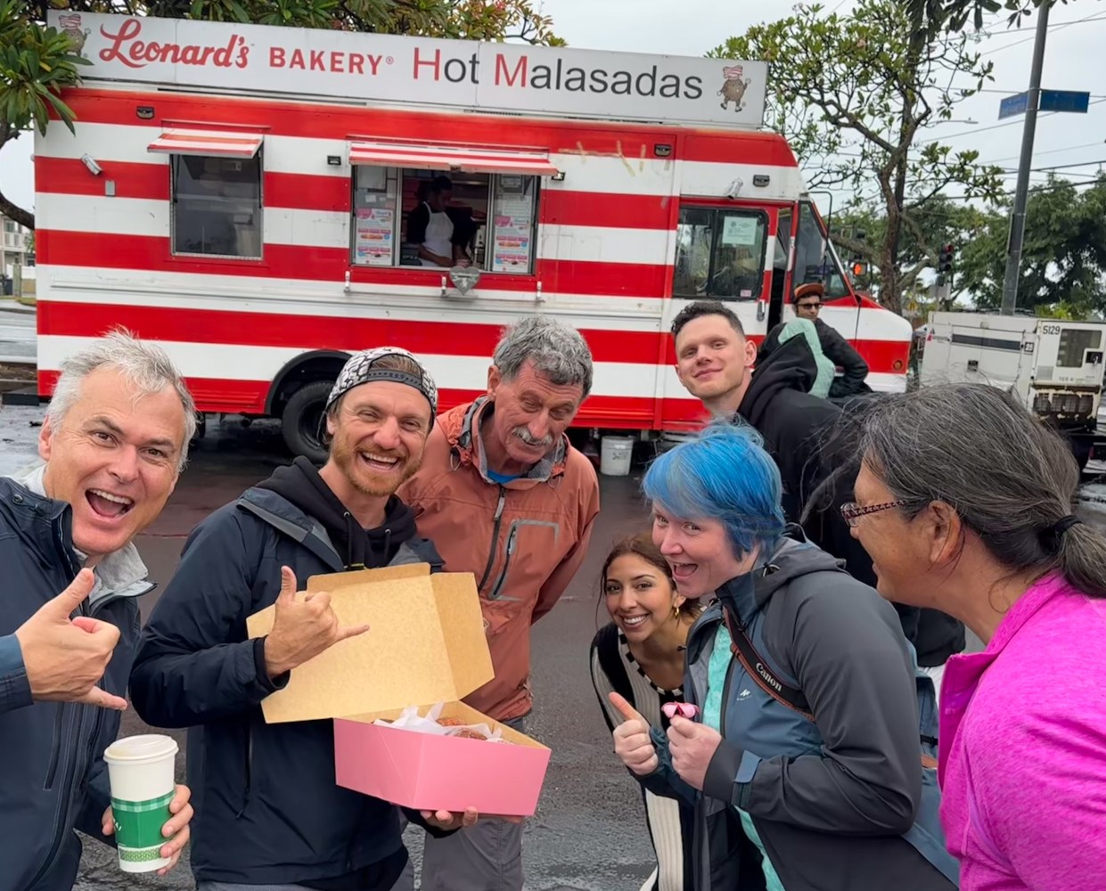 A group of happy tourists posing in front of the Leonard’s Bakery Malasada food truck in Hawaii. They are smiling, holding a pink box of fresh malasadas, and making shaka hand gestures. The food truck has red and white stripes, with a sign that reads 'Hot Malasadas.